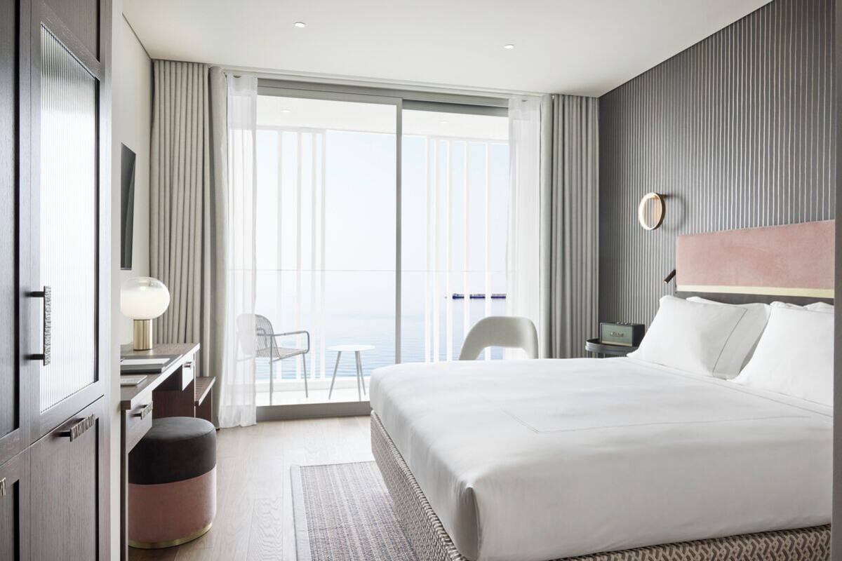 Une chambre d’hôtel avec un grand lit, un bureau avec une lampe, une chaise, un petit pouf et une vue sur la mer, accessible par une porte coulissante.