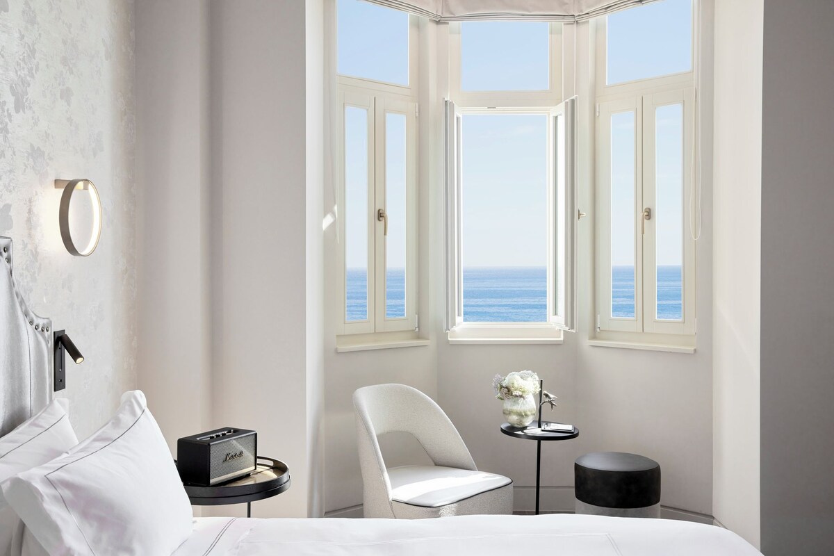A bedroom with a large window overlooking the sea, a bed, a chair, a small table with a vase, and a radio.