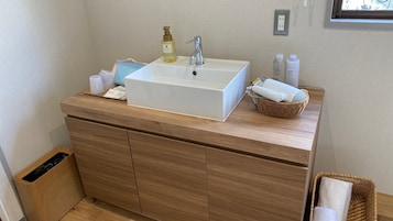 Un baño con lavamanos blanco, tocador de madera y una cesta de toallas.