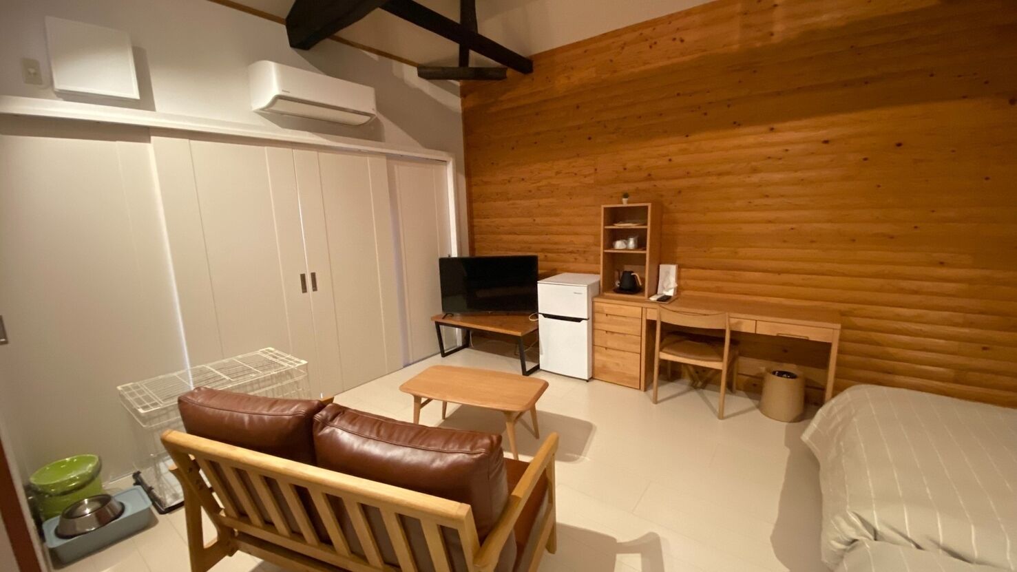 A compact room with a bed, a wooden desk, a television, and a pet feeding station.