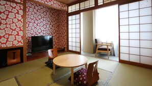A traditional Japanese room with tatami flooring, a tatami mat table, a television, and a sliding door leading to a balcony.