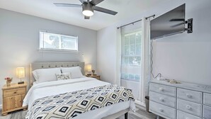 Un dormitorio con cama, mesitas de noche, cómoda, ventilador de techo y televisor montado en la pared.