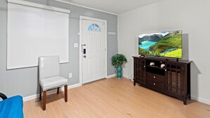Ein Zimmer mit einer weißen Tür, einem weißen Stuhl, Holzboden, einer dunklen Holz-Unterhaltungseinrichtung und einem Fernseher, der eine idyllische Landschaft zeigt.