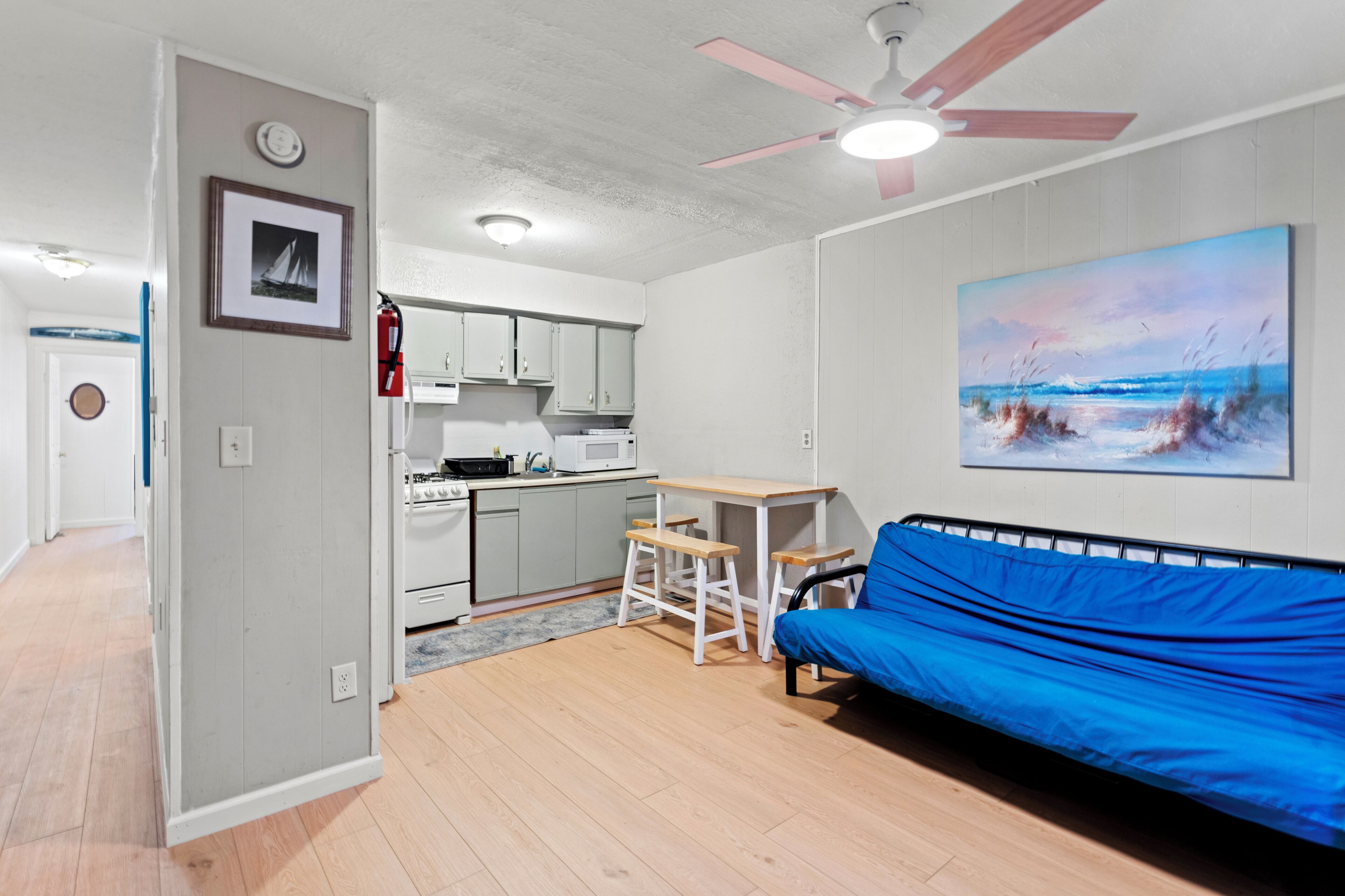 Un espace de vie compact comprenant un coin cuisine, un ventilateur de plafond, un canapé et un tableau représentant une scène de plage.