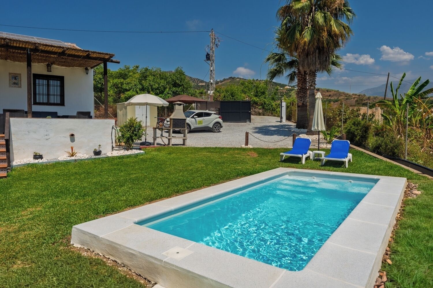 A rectangular swimming pool with a clear blue water surface, surrounded by a concrete deck. There are two blue lounge chairs and a small covered seating area with a table and chairs.