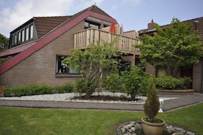 Una casa moderna con techo inclinado, terraza de madera y jardín con diversas plantas y un árbol en maceta.