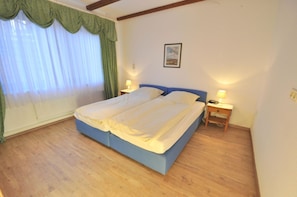 Una cama doble con cabecera azul en una habitación con piso de madera, dos mesitas de noche con lámparas y una ventana con cortinas traslúcidas.