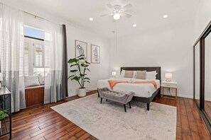 A bedroom with a bed, bedside tables, a nightstand, a ceiling fan, a potted plant, and a window with curtains.
