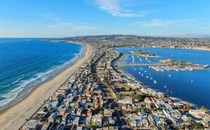 An aerial view of a coastal town with a long beachfront, a marina with boats, and dense residential buildings.