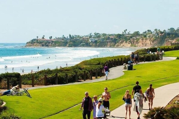 A coastal walkway with people walking, a grassy area, and a view of the ocean and hills in the background.