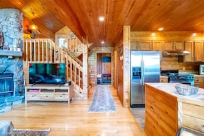 Interior de una cabaña de troncos con chimenea de piedra, escalera de madera, cocina con electrodomésticos de acero inoxidable y televisor en la sala de estar.