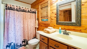 A bathroom with a wooden wall, a white sink, a mirror, and a shower curtain with a forest design. - Bear Ridge - Valley Views, Hot Tub, Dog Friendly (Davis)