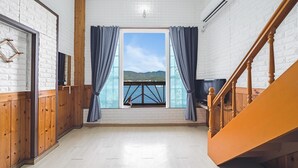 Una habitación con piso de madera, paredes de ladrillo blanco, un ventanal con cortinas, una escalera de madera y un televisor sobre un pedestal de madera.