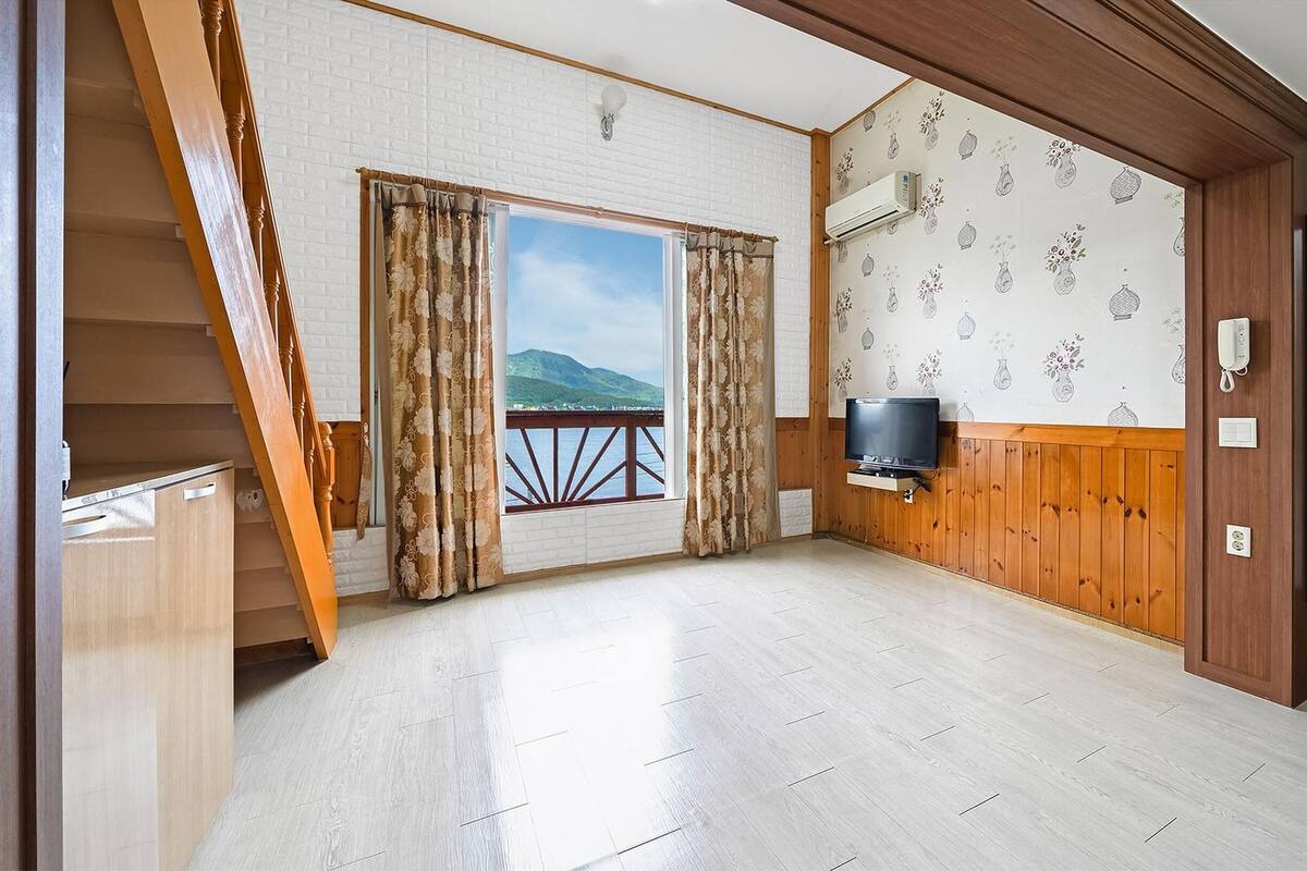 Una habitación con escalera de madera, televisor montado en la pared y ventana con cortinas.