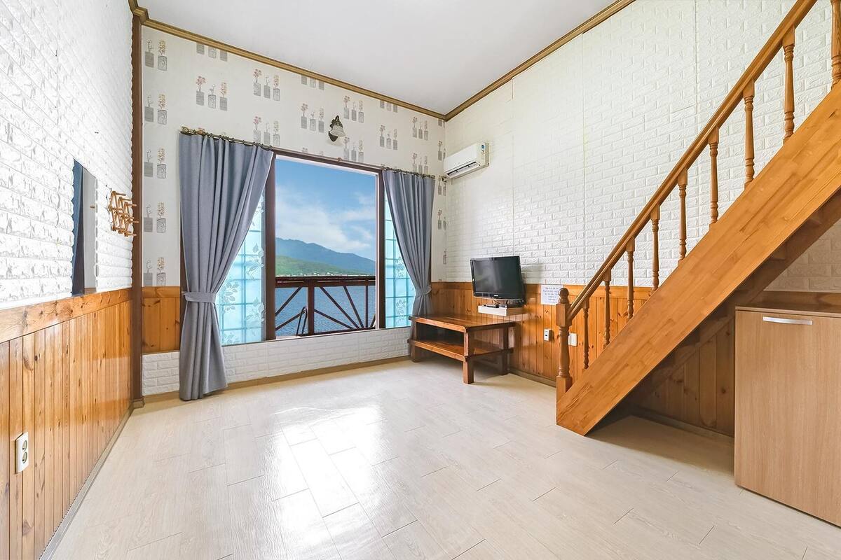 Una habitación con una escalera de madera, un televisor sobre un mueble bajo y una ventana con vistas a montañas.