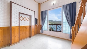 Una habitación con suelo de madera, paredes de ladrillo blanco, un ventanal con cortinas azules y vista al mar.