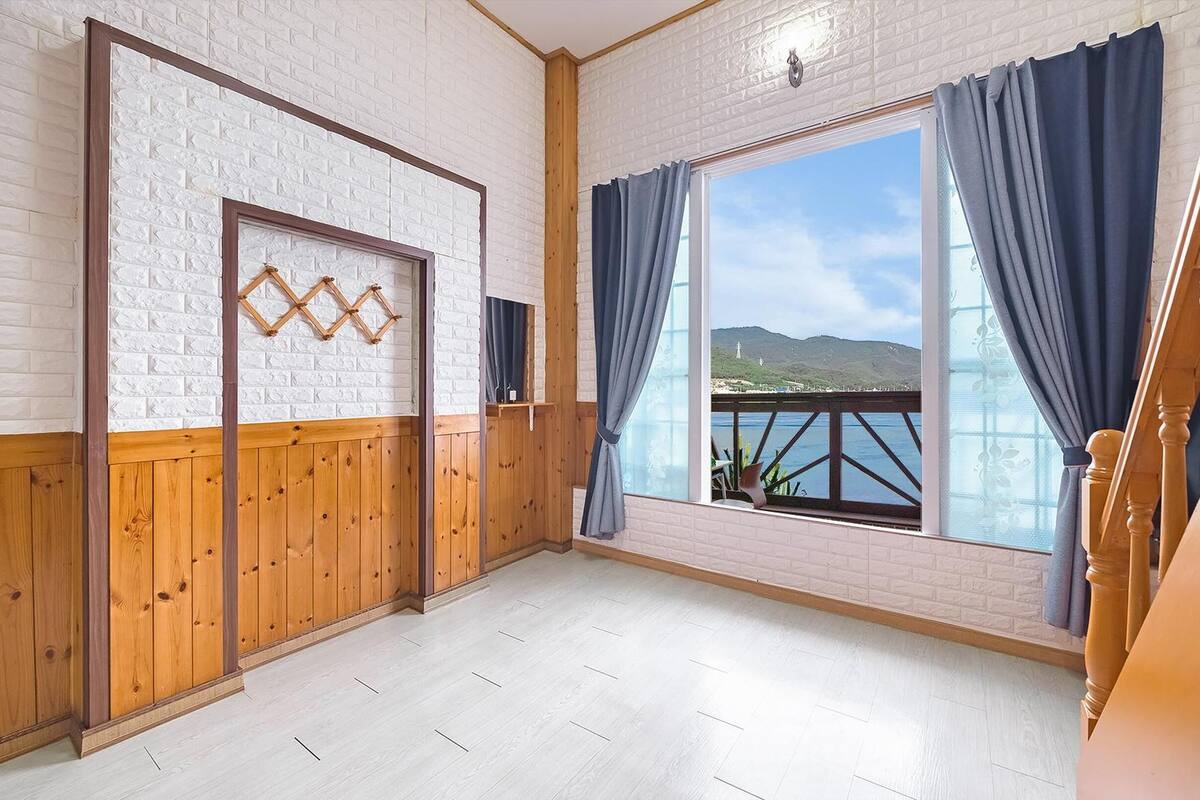 Una habitación con suelo de madera, paredes de ladrillo blanco, un ventanal con cortinas azules y vista al mar.