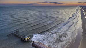 Vista aérea de una playa con un muelle que se adentra en el agua, olas suaves lamiendo la orilla y un cielo despejado al fondo.