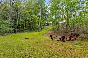 A cabin surrounded by dense forest, with a grassy area and Adirondack chairs near a fire pit.