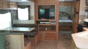 Un intérieur de camping-car compact comprenant un coin cuisine, un téléviseur fixé sur un meuble en bois et un lit avec une literie à motifs floraux.