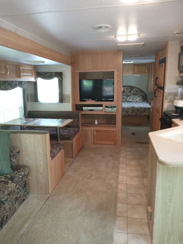 Ein kompaktes Wohnmobil-Interieur mit einer Küchenzeile, einem an einem Holzkasten montierten Fernseher und einem Bett mit blumigem Bettbezug.