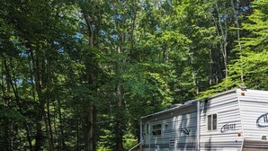 Un véhicule récréatif (VR) est stationné dans une zone boisée, sur un sol recouvert de gravier, à proximité d’un foyer et de chaises de camping.