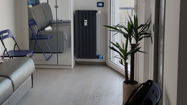 Una sala moderna con un sofá gris, una butaca azul, un radiador, una planta en maceta y un ventanal corredero que da a otra habitación.