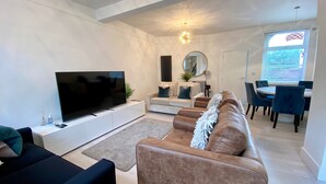 A modern living room with a flat-screen TV, leather sofas, a dining area with blue chairs, and a large window with blinds.