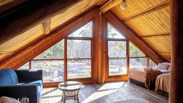 A cozy attic bedroom with a sloped ceiling, large windows, a bed, a chair, and a small table. - Girraween Country Inn (Eukey)