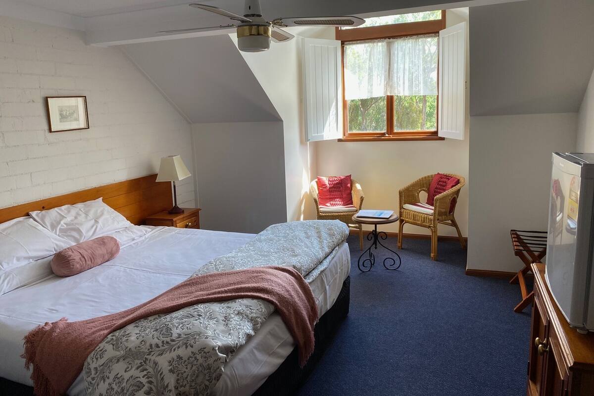 Un dormitorio con cama, ventilador de techo, ventana con cortinas, una silla, una mesita y un refrigerador.