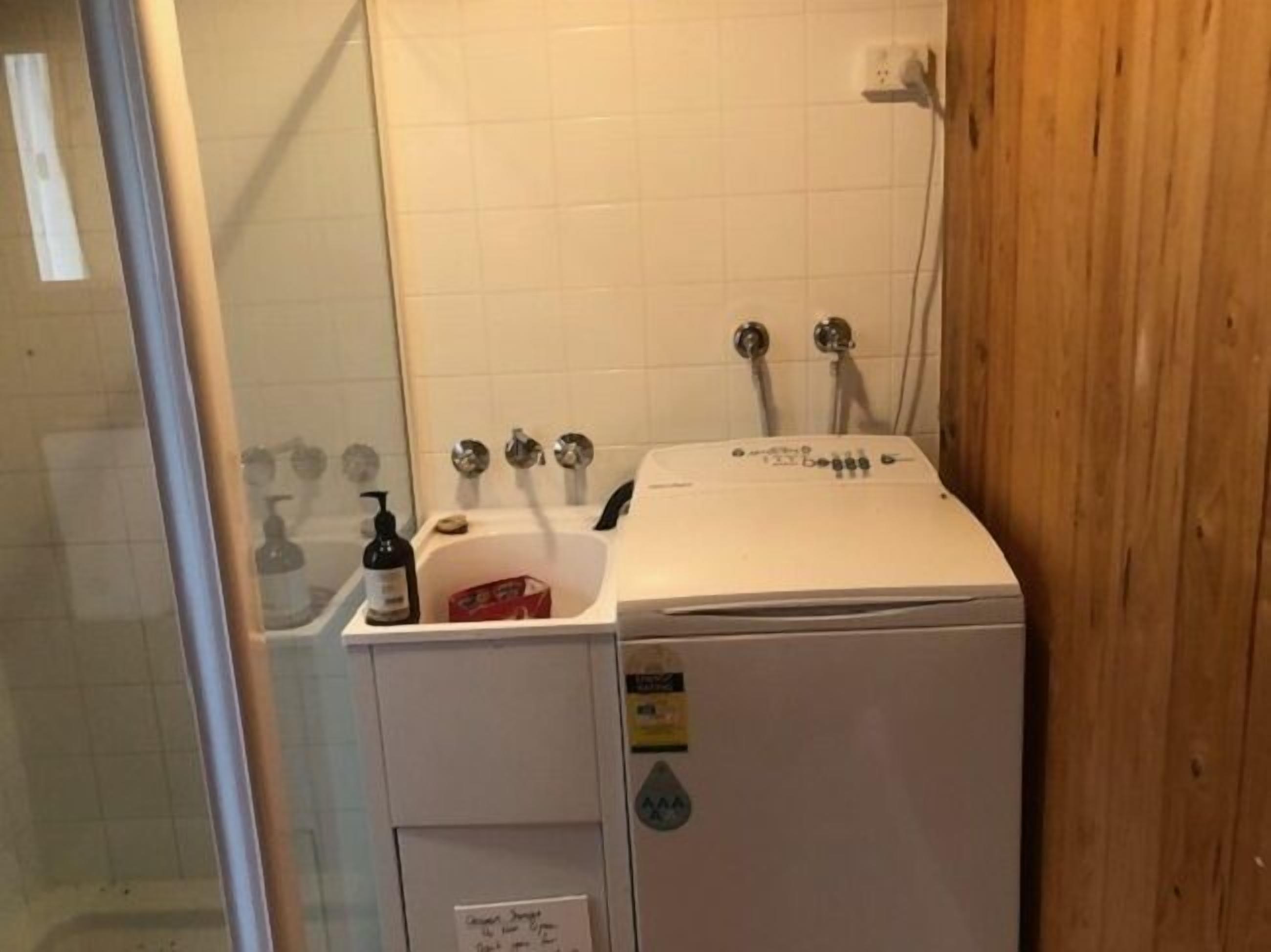 A compact, white laundry room with a washing machine, a small sink, and a shower area.