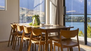 A dining area with a large table and chairs, a potted plant, and a view of mountains and a lake through large windows. - Fernlea by MajorDomo | Beautifully Renovated Home with Dramatic Lake Views, Spa Pool & Wrap-Around Veranda, Walk to Town (Queenstown)