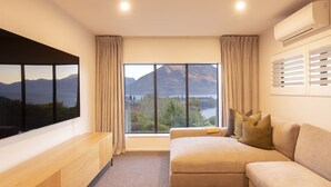 A modern living room with a large window, a sofa, a TV, and a wooden console. - Fernlea by MajorDomo | Beautifully Renovated Home with Dramatic Lake Views, Spa Pool & Wrap-Around Veranda, Walk to Town (Queenstown)