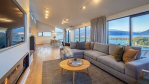 A modern living room with a large sofa, a coffee table, and a fireplace. There are large windows offering a view of a lake and mountains. - Fernlea by MajorDomo | Beautifully Renovated Home with Dramatic Lake Views, Spa Pool & Wrap-Around Veranda, Walk to Town (Queenstown)