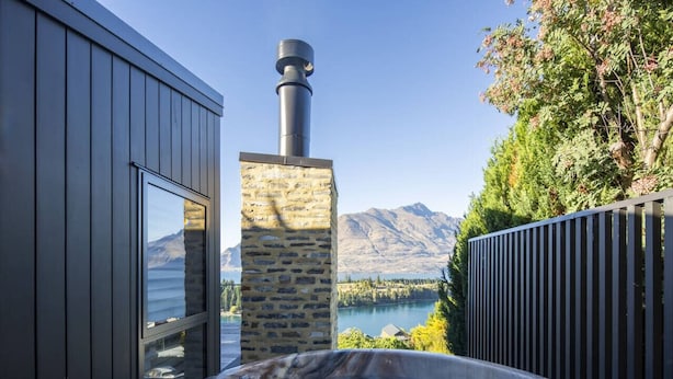 A hot tub with a stone chimney, a wooden deck, and a view of mountains and a lake. - Fernlea by MajorDomo | Beautifully Renovated Home with Dramatic Lake Views, Spa Pool & Wrap-Around Veranda, Walk to Town (Queenstown)