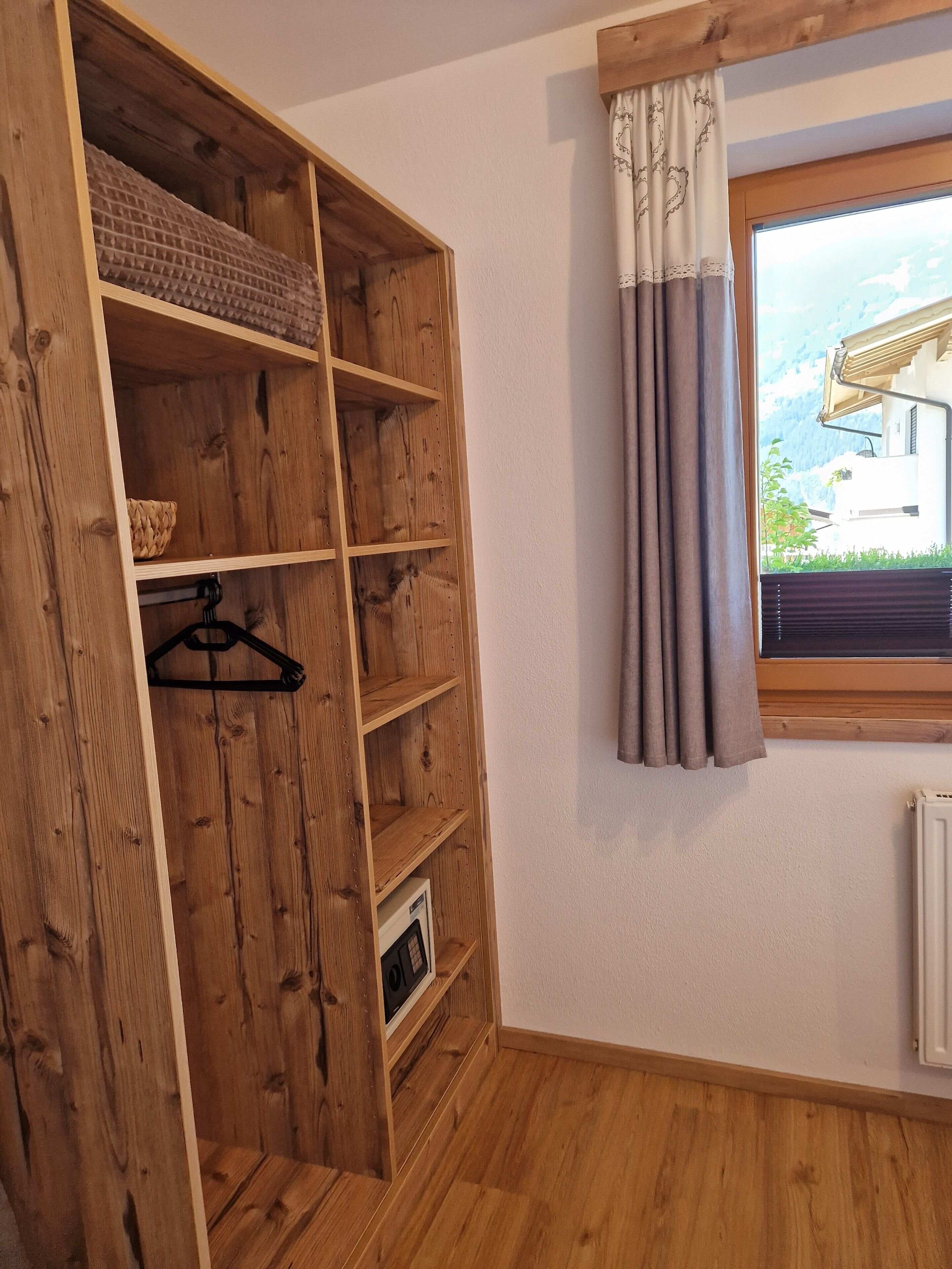 Un armario de madera con varios estantes y barra para colgar, un radiador y una ventana con cortinas.