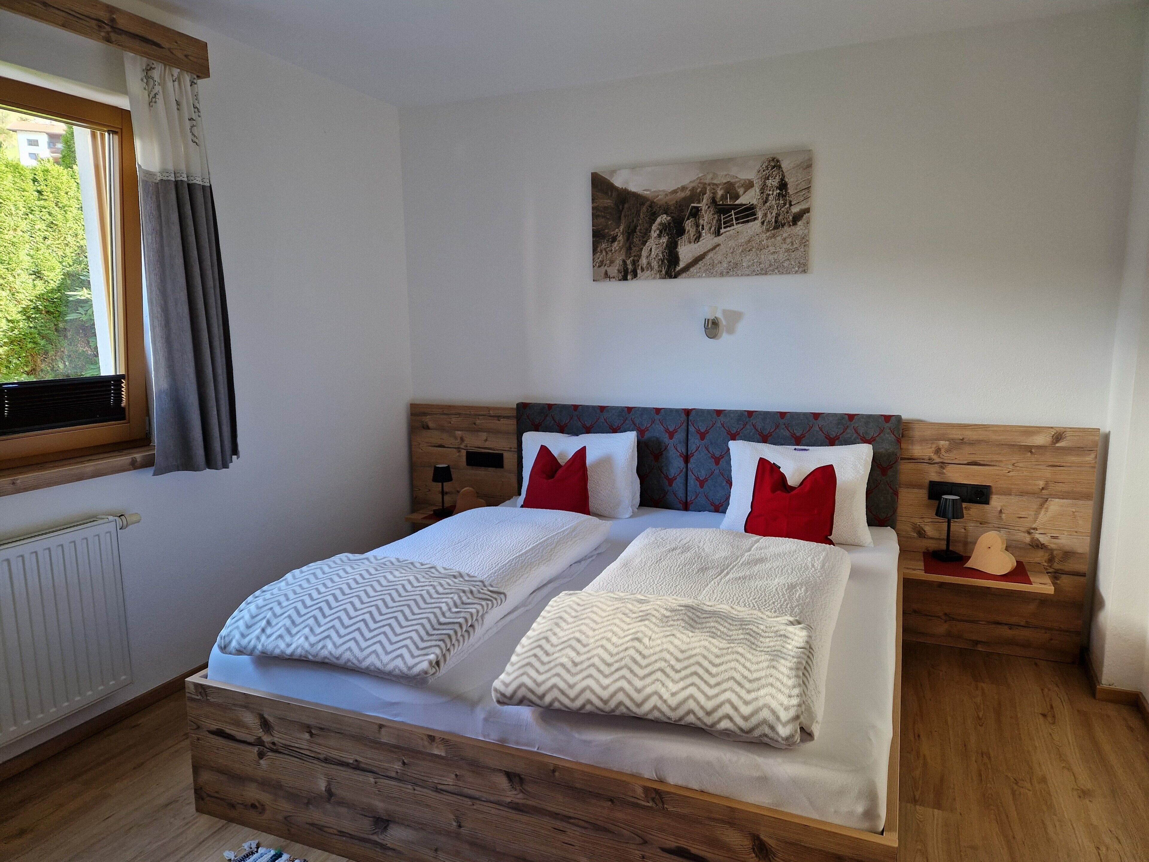 Une chambre avec une tête de lit en bois, un lit avec une literie blanche et des oreillers rouges, une fenêtre avec des rideaux, un radiateur et un tableau accroché au mur.