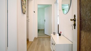 Un couloir avec un mur blanc, un plancher en bois, une commode blanche avec un miroir et une porte dotée d’une poignée en laiton.