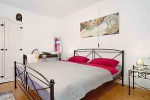 Un dormitorio con una cama, un armario, una mesita de noche con lámpara, un cuadro en la pared y una ventana con cortinas.