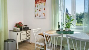 Un coin repas avec une table en bois, des chaises blanches, un comptoir de cuisine avec une machine à café et une affiche de cocktails accrochée au mur.