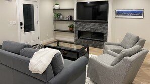 Ein modernes Wohnzimmer mit grauem Sofa, grauen Sessel, einem Glastisch, einem Kamin und einem an der Wand befestigten Fernseher.