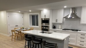 Eine moderne Küche mit weißen Schränken, einer großen Kochinsel mit Barhocker, einem eingebauten Backofen und einer Edelstahl-Dunstabzugshaube.