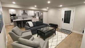 Ein modernes Wohnzimmer mit einem grauen Sofa, einem Glastisch und einer Küchenzeile im Hintergrund.