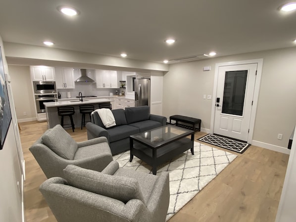 A modern living room with a grey sofa, a glass coffee table, and a kitchen area in the background.