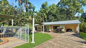 Un espacio al aire libre cercado con una zona de estar cubierta, un camino que lleva a un edificio y una variedad de árboles y plantas.