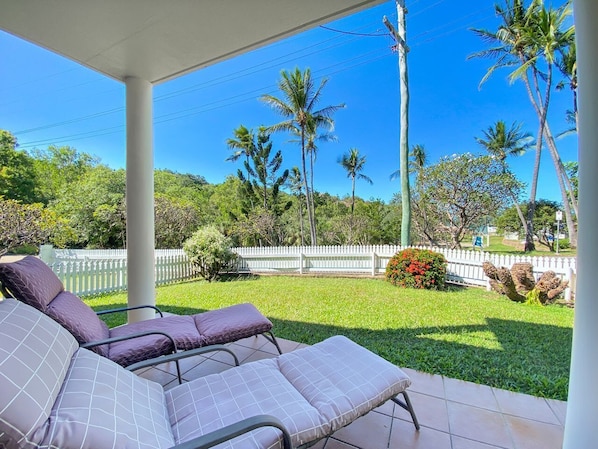 A covered patio with two lounge chairs, a white picket fence, and palm trees in the background.