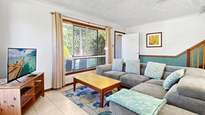 Una sala de estar con un sofá, una mesa de centro, un televisor sobre un soporte de madera y una ventana con cortinas.