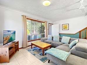 Ein Wohnzimmer mit einer Couch, einem Couchtisch, einem Fernseher auf einem Holzboden und einem Fenster mit Vorhängen.
