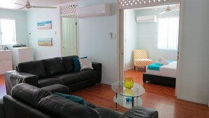 Ein Wohnzimmer mit einem schwarzen Sofa, einem Glastisch und einem Bett mit einer blau-weiß gestreiften Decke.