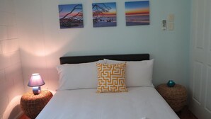 Ein Bett mit weißer Bettwäsche, einem gemusterten Kissen, einer Lampe auf einem Korbstuhl-Nachttisch und drei gerahmten Bildern an der Wand.